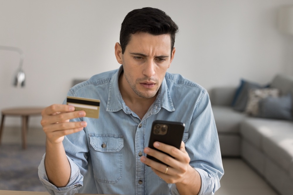 Concerned man checks his phone while holding a credit card after a missed payment