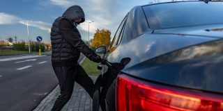 Person in a hooded jacket and face covering attempting to break into a parked black car by pulling on the driver's side door handle, illustrating vehicle theft and anti-theft car insurance.