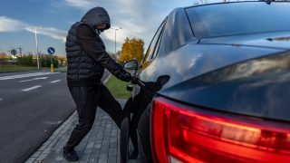 Person in a hooded jacket and face covering attempting to break into a parked black car by pulling on the driver's side door handle, illustrating vehicle theft and anti-theft car insurance.