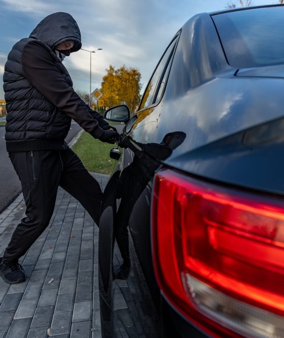Person in a hooded jacket and face covering attempting to break into a parked black car by pulling on the driver's side door handle, illustrating vehicle theft and anti-theft car insurance.