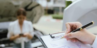 Insurance agent completing a car insurance quote on a clipboard beside a vehicle with its hood open while a driver stands nearby using a phone.
