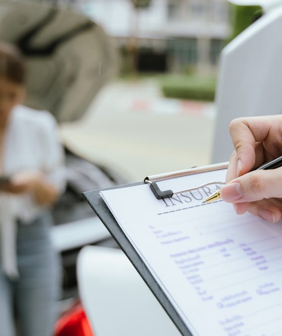 Insurance agent completing a car insurance quote on a clipboard beside a vehicle with its hood open while a driver stands nearby using a phone.
