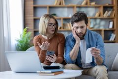 A concerned couple reviews rising household expenses together, looking at a bill, smartphone, and laptop while discussing the impact of home inflation.