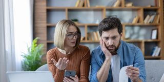 A concerned couple reviews rising household expenses together, looking at a bill, smartphone, and laptop while discussing the impact of home inflation.