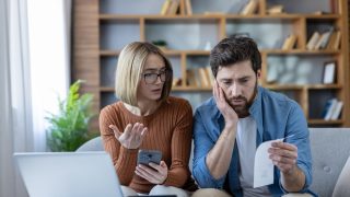 A concerned couple reviews rising household expenses together, looking at a bill, smartphone, and laptop while discussing the impact of home inflation.