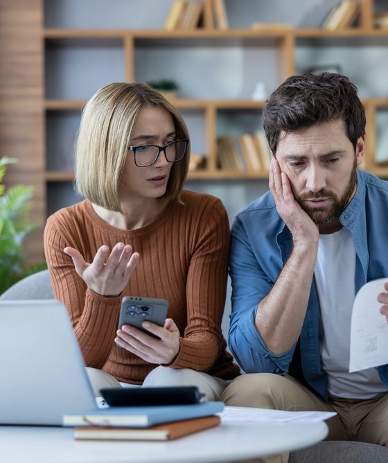 A concerned couple reviews rising household expenses together, looking at a bill, smartphone, and laptop while discussing the impact of home inflation.