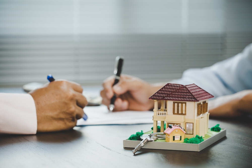 A model house and keys sit on a table as two people review and sign housing documents in the background.