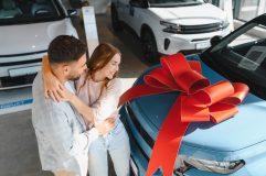 Couple admiring a new blue car with a large red bow at a dealership while shopping for new car insurance.