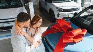 Couple admiring a new blue car with a large red bow at a dealership while shopping for new car insurance.