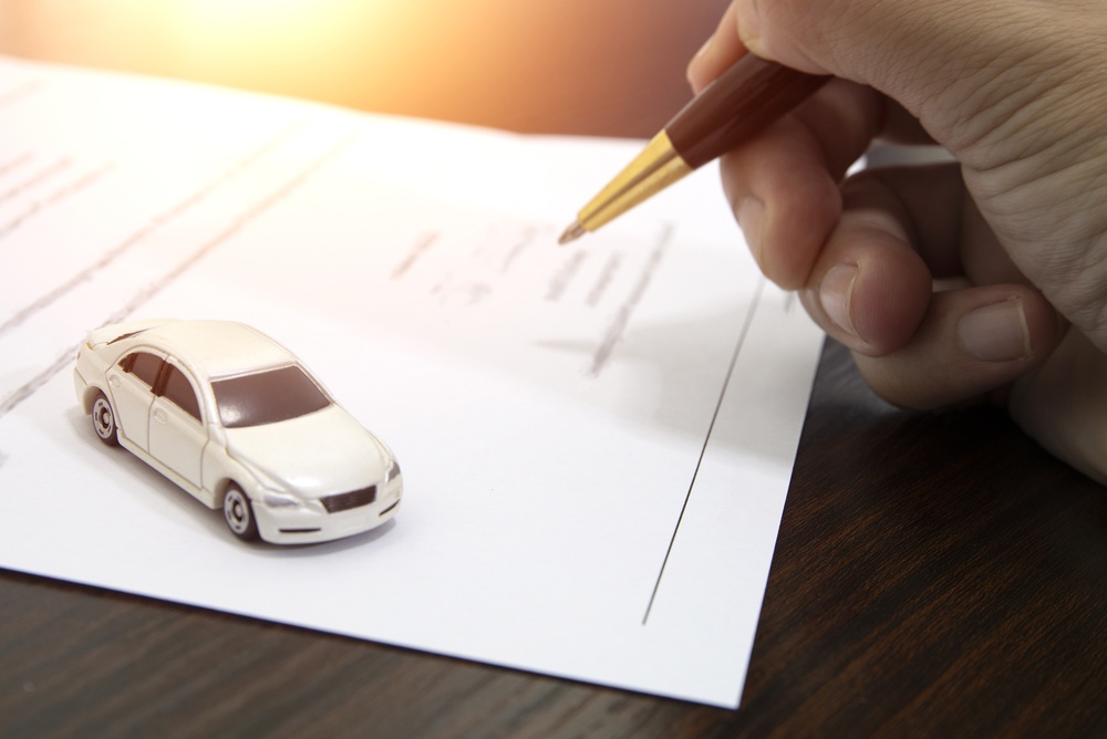Hand holding a pen over car insurance paperwork beside a miniature white car, representing new car insurance coverage.