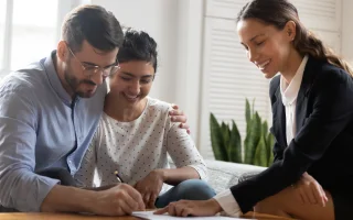 Husband in glasses embracing wife, putting signature on car insurance documents with friendly insurance agent, cheap car insurance.