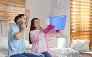 Young couple wife holding bucket with leak coming in the bedroom husband on phone agitated leaking roof.