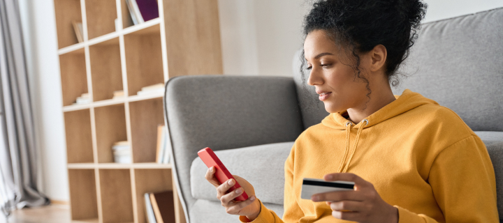 Young adult African American female consumer holding credit card and smartphone sitting on floor at home doing online banking transaction. E commerce virtual shopping, secure mobile banking concept.