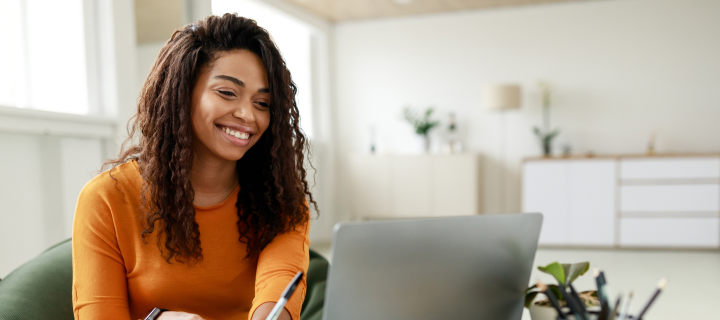 Smiling young African American woman sitting at desk working on laptop taking notes in notebook, happy millennial female studying online, watching webinar using computer and writing check list.