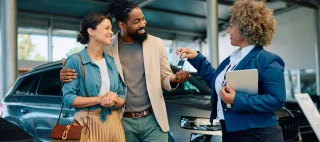 Happy multiracial couple receiving keys of their new car from saleswoman in showroom – cheap car insurance.