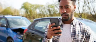 Young man standing by damaged car after traffic accident calling breakdown recovery company using mobile phone – cheap car insurance.