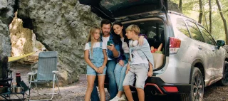 Happy family with their son and daughter using phone sitting in trunk of car while traveling during summer vacation road trip. Pretty family mother father and children sitting in car with trunk open.