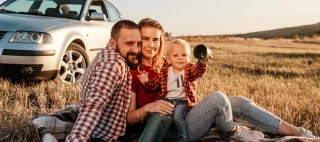Happy Young Family Mom and Dad with Their Little Son Enjoying Summer Weekend Picnic Sitting on the Plaid Near the Car Outside the City in the Field.
