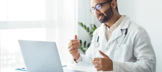 Portrait of indian man doctor talking to online patient on laptop screen sitting at clinic office desk giving online consultation for domestic health treatment. Telemedicine remote medical appointment.