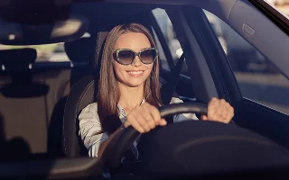 Photo of charming sweet young lady dressed white shirt dark eyewear smiling driving automobile outdoors urban route.
