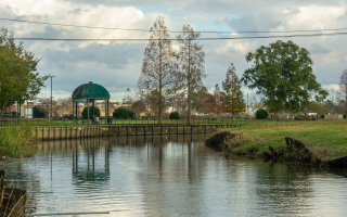 A calm pond in Jambalaya Park in Gonzales, Louisiana, with a small green-roof gazebo by the water, trees along the shoreline, and a cloudy sky reflected in the water.