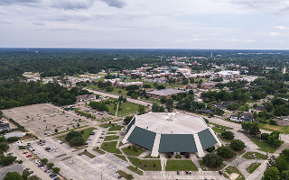 A wide aerial drone photograph of the campus of Southeastern Louisiana University in Hammond, Louisiana, showing the distinctive octagonal arena or auditorium building prominently in the mid-foreground, with the broader SLU campus spreading into the background and the suburban and forested landscape of Hammond extending to the horizon.