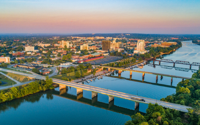 Aerial view of downtown Augusta, Georgia, with the Savannah River in the foreground, multiple bridges crossing the river, mid-rise buildings, and tree-lined riverbanks at sunset.