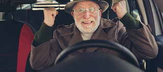 An old pensioner raises fists, a toothy smile, wears a brown jacket, headwear, and eyeglasses inside a car.